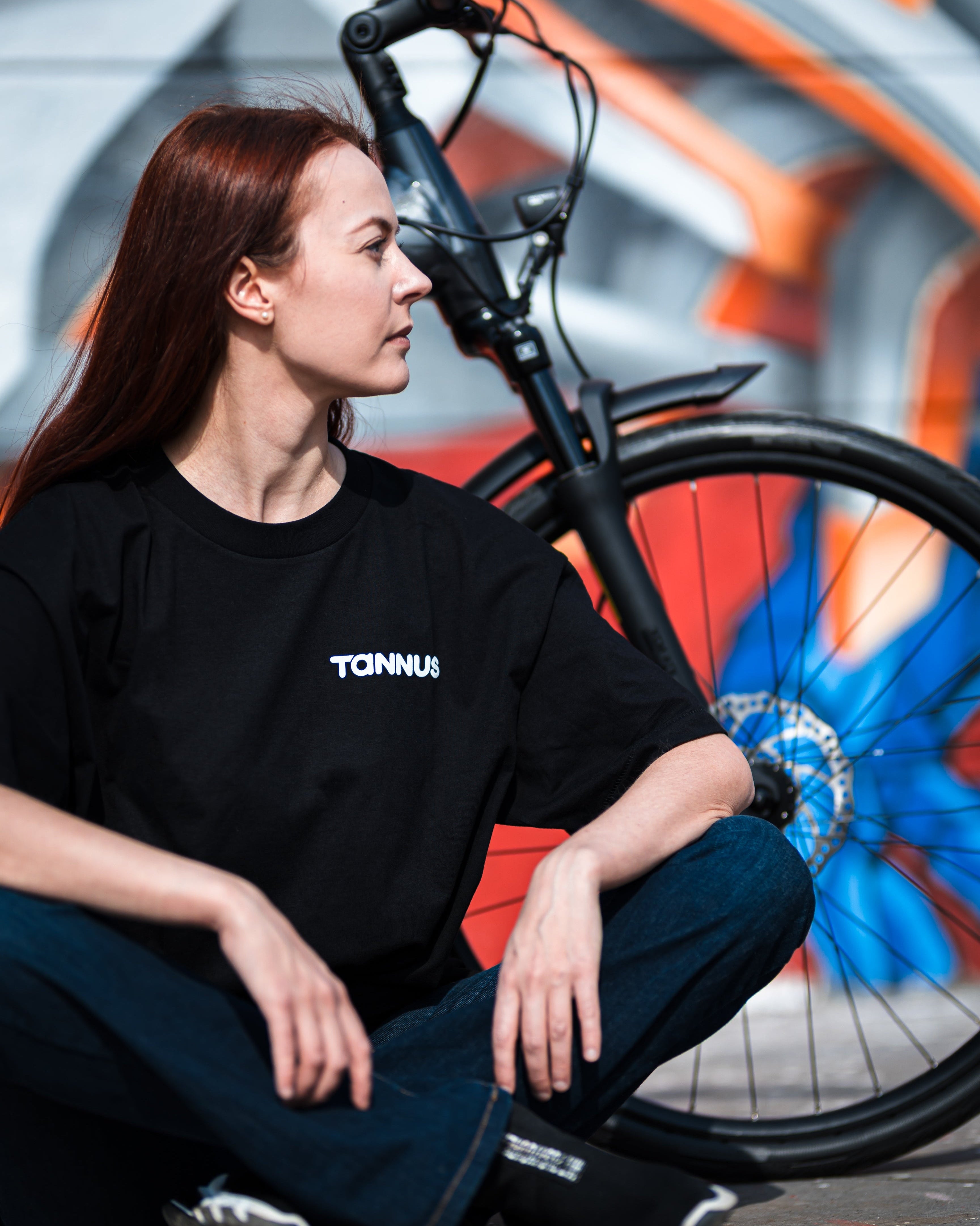 Person wearing a black TANNUS shirt sitting next to a bicycle with colorful abstract art in the background.